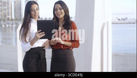 Two stylish attractive young women on a waterfront esplanade standing chatting and looking at a tablet computer together Stock Photo