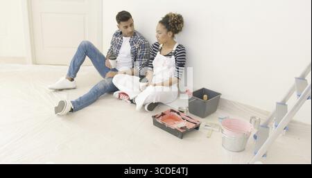 Young couple taking a break from painting their apartment sit on the floor with cups of takeaway coffee having a serious discussion Stock Photo