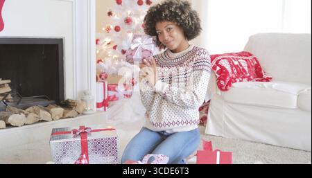 Pretty happy young woman checking her Christmas gifts on the floor in her living room holding up a decorative box tied with a bow with friendly smile Stock Photo