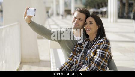 Happy laughing couple taking their selfie on a smartphone as they pose close together on a sunny urban promenade Stock Photo