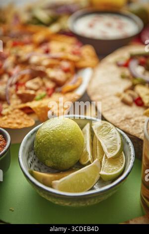 Close up on lime slices in ceramic bowl with various freshly made Mexican foods assortment in the back. Placed on colorful table. With nachos, tacos, Stock Photo