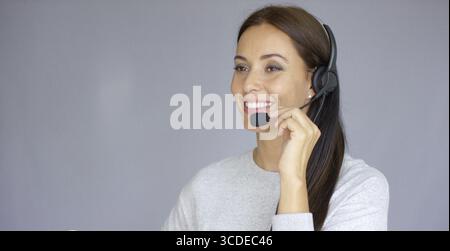 Beautiful and positive female call center agent at work. She speaking with customer using headset. Smiling. Isolated on gray background Stock Photo
