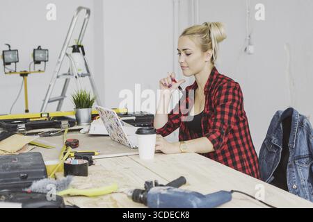 Beautiful young woman in checkered shirt sitting at wooden workbench with carpenter tools and working on laptop smiling Stock Photo