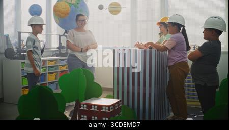 Team of Primary School Students in Hardhats Discussing City Planning with Large Cardboard Urban Blocks and Models of Trees, Buildings. Female Teacher Organizing Interactive STEM Class. Dolly Shot. Stock Photo
