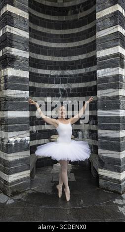 Ballerina in a white dress dancing in front of black and white striped architecture, San Giovanni Battista, Mogno, Ticino, Switzerland Stock Photo