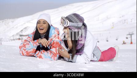 Cute pair of female twins in ski clothing laughing while laying down on ground at bottom of ski slope with mountain behind them Stock Photo