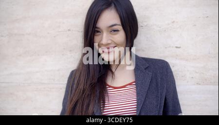 Gorgeous young woman with long brunette hair and a vivacious smile looking at the camera head and shoulders against a texture wall with copy space Stock Photo
