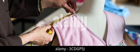 Caucasian woman measuring shoulder seam of dress on mannequin using measuring tape, hands adjusting fabric for precise tailoring in sewing workshop environment Stock Photo