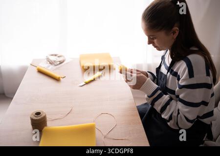 Hispanic woman in apron making beeswax candles using natural wax sheet. Side view. Close-up Stock Photo