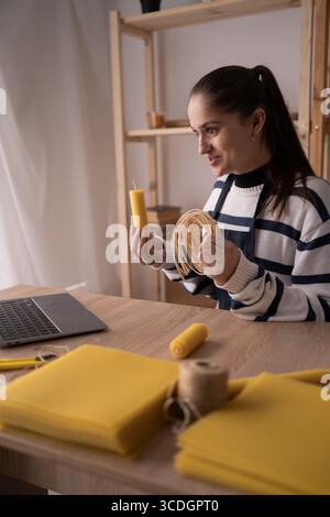 Woman creating boutique candles from beeswax at home, working on laptop at table, creative entrepreneur lifestyle, diy handmade product concept Stock Photo