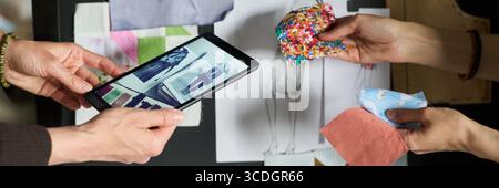 Caucasian woman holding tablet displaying fashion sketches while another Caucasian woman selecting colorful fabric swatches, collaborating on clothing design project at workspace Stock Photo