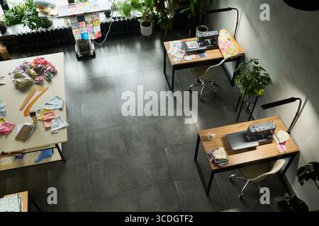 Empty sewing studio showing two workstations with sewing machines and fabric pieces on desks, cutting table with fabric scraps and sewing tools, indoor plants decorating workspace Stock Photo
