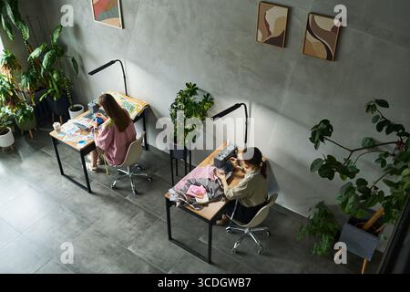 Two young adult Caucasian women sitting at separate desks working on creative projects, one drawing with colored pencils, other using sewing machine, surrounded by indoor plants Stock Photo