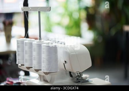 Industrial overlock sewing machine standing on table with multiple large thread spools attached, threads running through guides, blurred workspace background visible Stock Photo