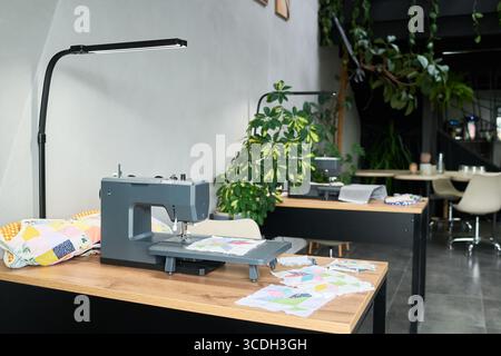 Modern sewing studio featuring sewing machine on wooden table with colorful patchwork fabric, bright workspace surrounded by green plants, empty chairs in background Stock Photo