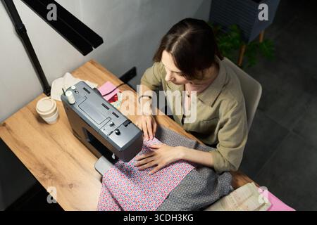 Young adult Caucasian woman using sewing machine, focusing on stitching patterned fabric at wooden desk, hands guiding textile, working in creative workspace, top view perspective Stock Photo