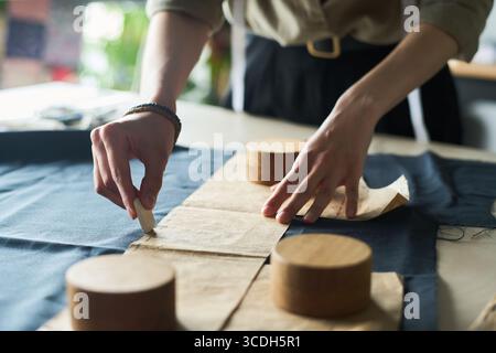Caucasian young adult woman marking fabric with tailors chalk while preparing sewing pattern, hands visible working on textile project, wooden weights holding pattern pieces in place Stock Photo