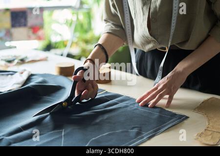 Caucasian young adult woman cutting fabric with large scissors on worktable, measuring tape around neck, hands carefully guiding textile, engaged in garment making process Stock Photo