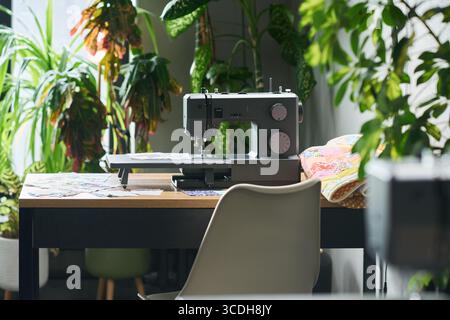 Sewing machine positioned on desk surrounded by green plants, fabric pieces scattered on tabletop, empty chair placed in front, natural light illuminating workspace Stock Photo