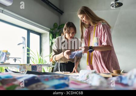 Two young adult Caucasian women collaborating on textile project, examining fabric samples together in bright workspace, sewing tools and materials visible on table in foreground Stock Photo