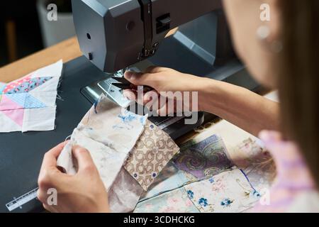 Young adult Caucasian woman sewing fabric pieces with sewing machine, hands adjusting textile while working on patchwork quilt, close up of creative process in crafting environment Stock Photo