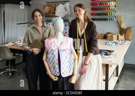 Portrait of two Caucasian young adult women standing in sewing studio smiling at camera, one holding tablet, mannequin dressed in colorful patchwork garment between them, spools of thread on wall Stock Photo