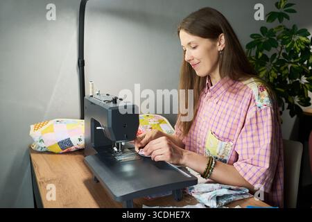 Caucasian young adult woman using sewing machine, creating patchwork quilt at wooden table, smiling and focusing on fabric, hands guiding material under needle, indoor setting Stock Photo