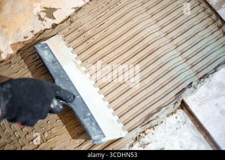 Close-up of notched trowel spreading tile adhesive evenly before laying ceramic floor tiles. Stock Photo
