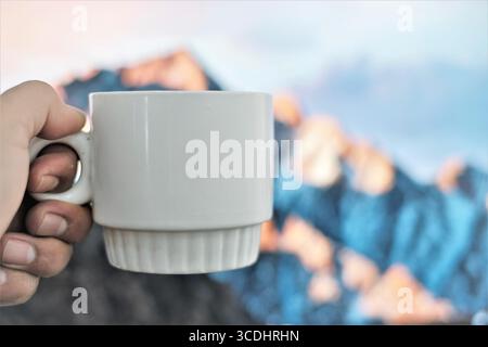 Man's hand holding a white cup, with a cliff image in the background. Stock Photo
