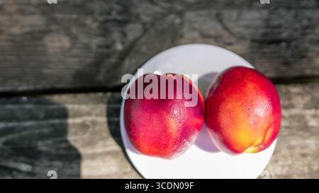 Two ripe nectarines with vibrant skin are placed on a white plate. The wooden table adds a rustic charm, while soft natural light enhances their color Stock Photo
