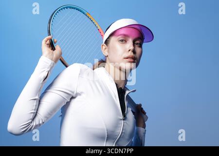 Young woman athlete in white sportswear holding racket on shoulder against blue studio background confident modern portrait for sport and lifestyle Stock Photo