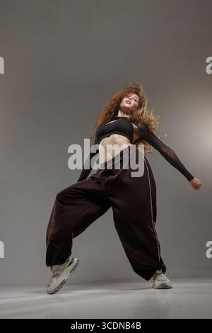 Young female dancer in stylish outfit performing energetic jazz funk move with attitude and expression in studio. Stock Photo