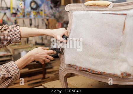 Female restorer using tools to repair vintage chair upholstery in workshop, showing detailed furniture restoration process Stock Photo
