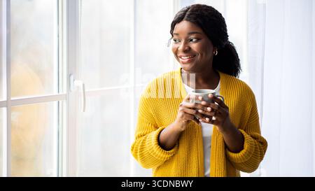 Black lady standing looking through window, drinking tea Stock Photo