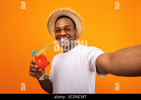 Good looking black guy in summer wear drinking yummy fruit cocktail and taking selfie on yellow studio background Stock Photo