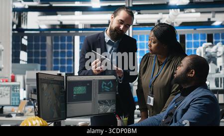 Multiethnic team of experts collaborating in a solar panel power plant, reviewing blueprints and production line metrics to ensure clean energy manufacturing and sustainability. Camera B. Stock Photo