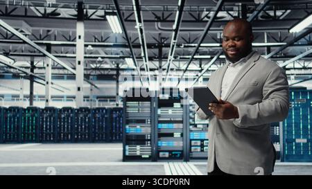 Cheerful data center programmer looking at code debugging tools running on tablet. Server farm IT professional typing on device touchscreen, writing script automating tasks, camera A Stock Photo