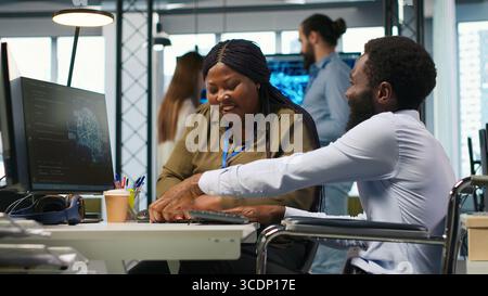 Startup office engineer in wheelchair talking with colleague, developing artificial intelligence models. IT specialist with paraplegia and coworker running simulations for AI forecasting, camera A Stock Photo