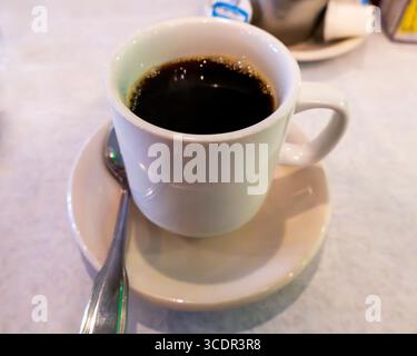 A steaming cup of black coffee sits on a white saucer with a spoon, ready to be enjoyed. The deep, rich liquid has a slight foam on top, inviting a wa Stock Photo