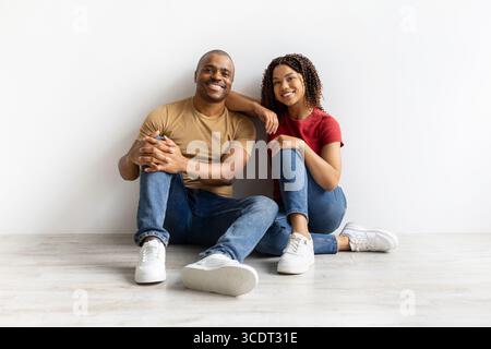 Relaxed black couple sitting on floor against white wall, smiling at camera Stock Photo