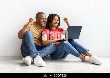 Excited black couple sitting on floor, using laptop together and celebrating success Stock Photo