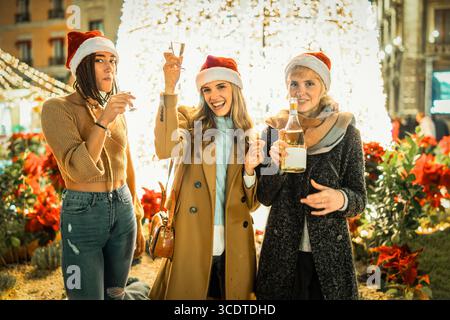 Happy multicultural group of three young female friends in Santa hats looking at the camera and toasting with champagne at an outdoor warm climate Chr Stock Photo
