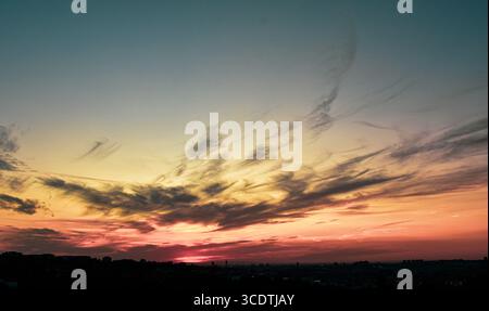 Hazy clouds fading from orange to grey above dark building silhouettes at sunset. Stock Photo
