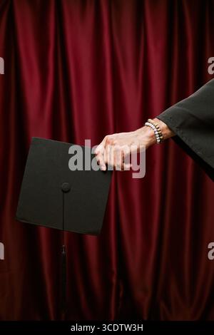 Hand of young adult man holding graduation cap against red curtain background, wearing bracelet and academic gown, celebrating achievement in photo booth setting Stock Photo