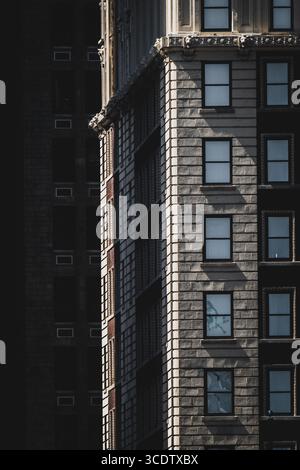 View of light playing on the textured facade of a tall building with evenly spaced windows, casting shadows on the adjacent dark structure, New York, New York, United States. Stock Photo