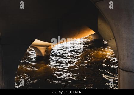 View of shimmering water reflecting golden light beneath the concrete arches, a stark contrast of textures and tones, creating a mesmerizing scene, Ne Stock Photo