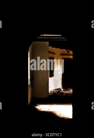 An interior shot of an abandoned building, with a dark foreground and a brightly lit hallway in the background. Debris litters the floor. Stock Photo