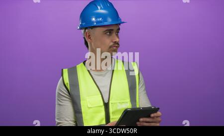 Young man in reflective vest and hardhat examines a tablet against isolated purple background, symbolizing technology and construction with a professi Stock Photo