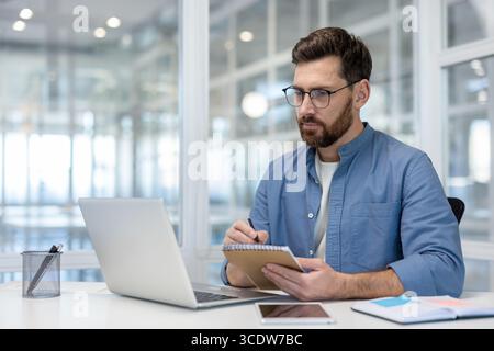 A man wearing glasses, using a laptop and taking notes while sitting in an office. The setting features a well-lit, modern design, promoting an atmosphere of concentration Stock Photo