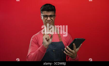 Young man wearing glasses and apron interacts with tablet against vibrant red background showcasing modern technology and casual style in a captivatin Stock Photo